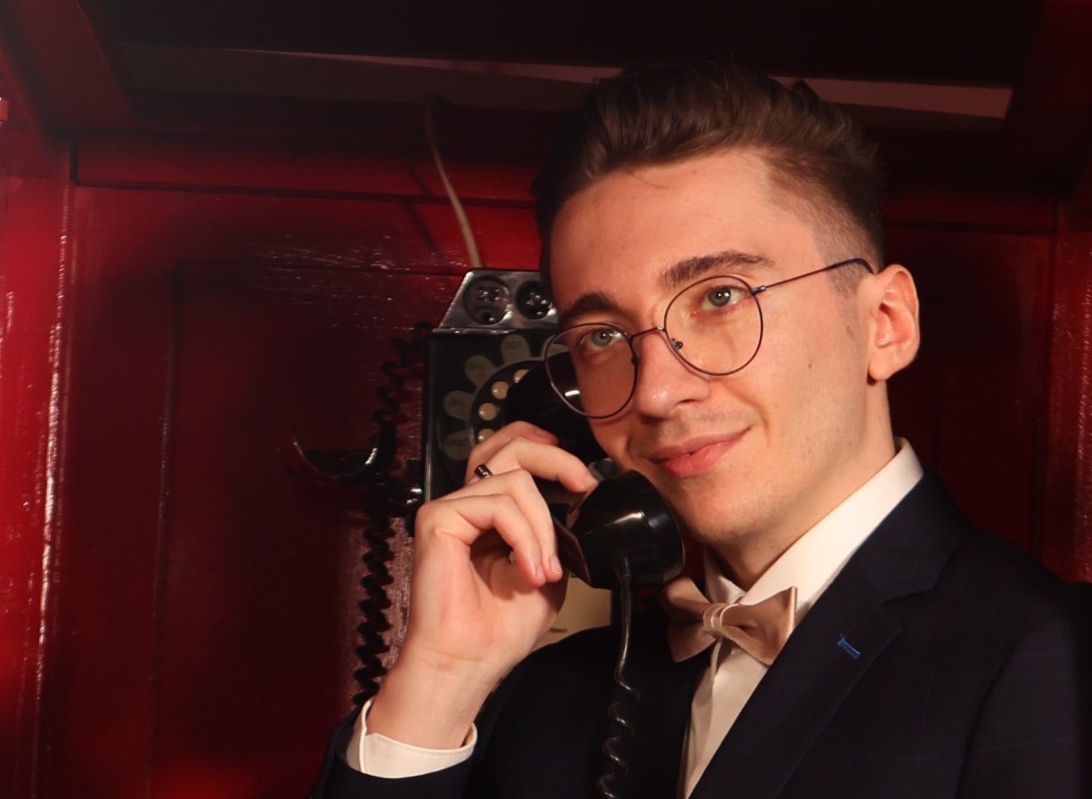 Remy Machado in a red phone booth, smiling, bow tie on, holding the receiver of a rotary phone